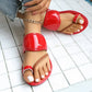 Women's red patent toe-thong flat sandals with a braided strap detail, shown on feet with white-painted toenails against a white tile background. A delicate gold ankle chain is visible.