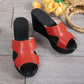 Pair of elegant red peep-toe wedge sandals with a single geometric strap, resting on a grey wooden floor with decorative yellow flowers and a woven basket.