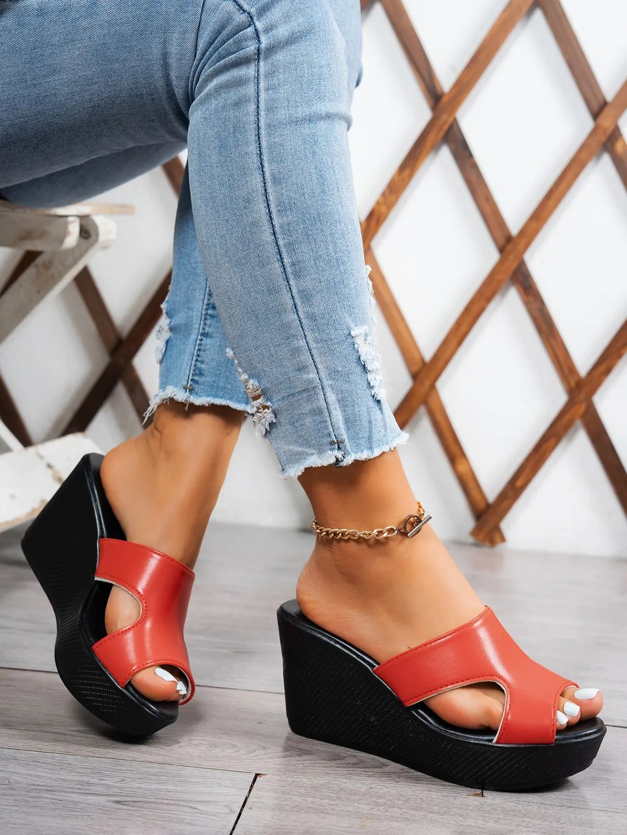 Elegant red peep-toe wedge sandals with a black textured sole and a single supportive strap, shown on a model's feet with ripped denim jeans and a gold anklet.