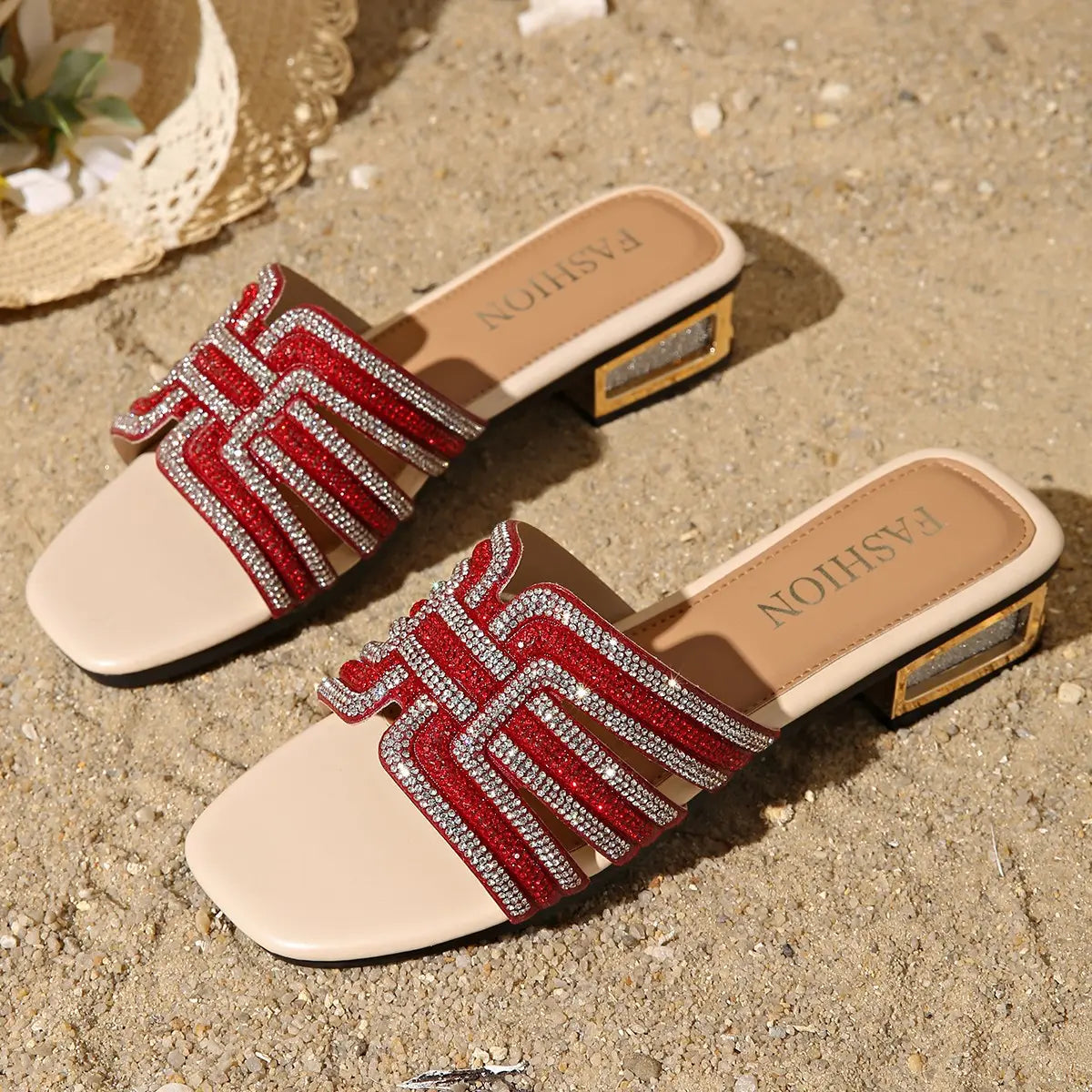 Red embellished flat slide sandals with geometric rhinestone detailing and a metallic glitter block heel, shown on sand with a straw hat accent.