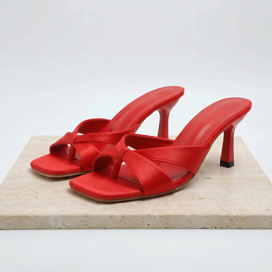 Red PU square-toe stiletto mules with a central knot detail and thong toe post, presented on a marble block against a white studio background. These elegant slip-on sandals feature a 7cm flared heel.