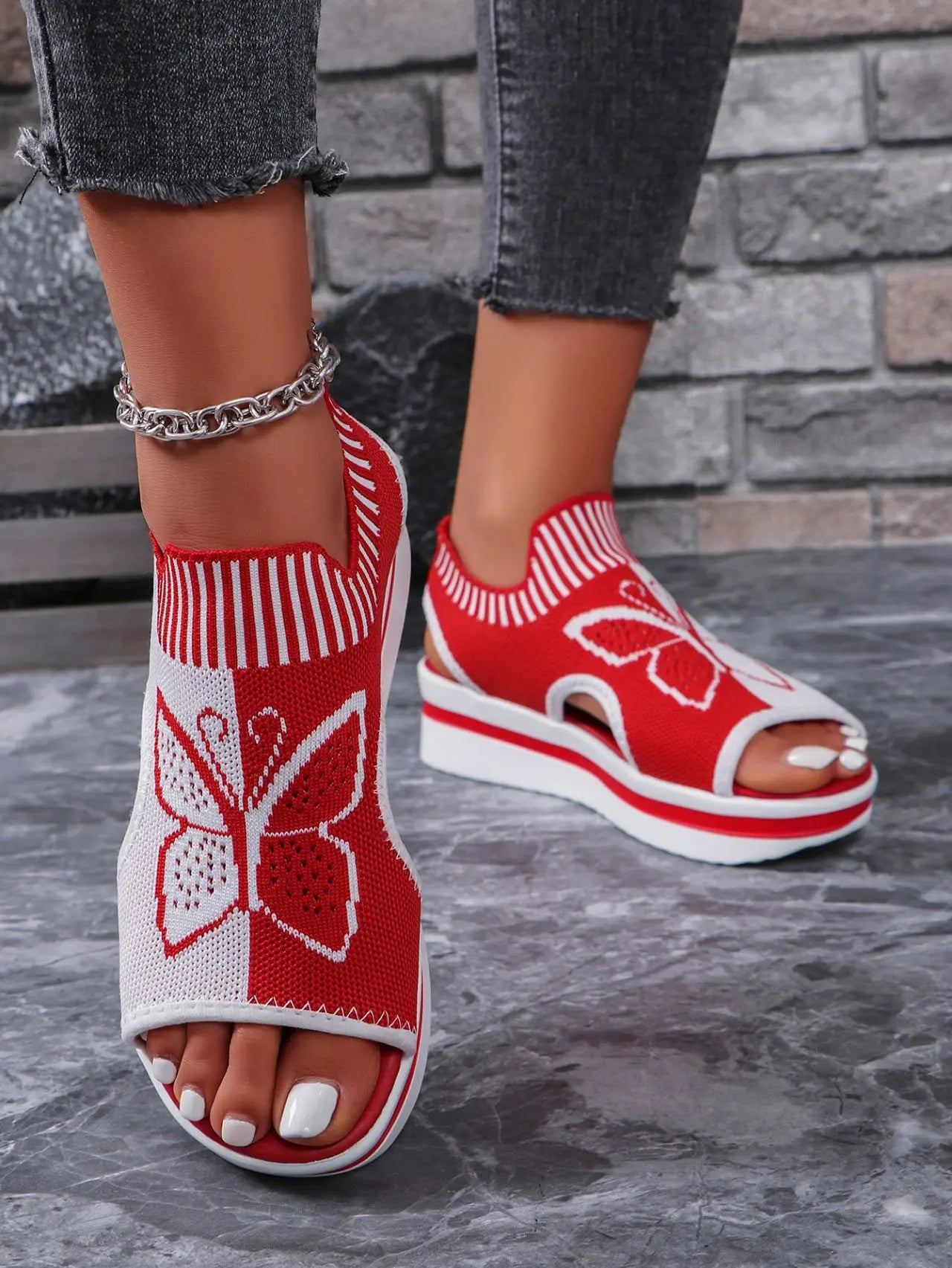 Red and white women's knit wedge sandals with a detailed butterfly motif, peep-toe design, and slingback strap, worn by a model with dark denim jeans.