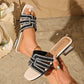 Elegant champagne shimmer embellished flat sandals worn by a model, showcasing intricate geometric rhinestone straps, a square toe, and a shimmering metallic block heel on a sandy beach setting.