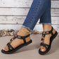 Black embellished flat Rhyko sandals with a T-strap featuring floral beadwork and a bow, shown on a model's foot with distressed denim jeans and an anklet, set against a rustic stone and wood plank background.