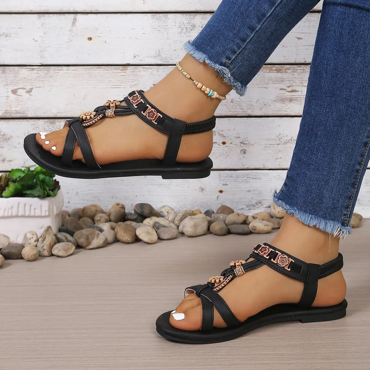 Black Rhyko boho chic flat sandals featuring intricate floral beadwork and a delicate bow on the T-strap, with a comfortable elasticated slingback strap worn on a woman's foot with blue denim jeans and a beaded anklet.