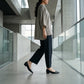 A woman walks forward in black Rhylo pointed-toe flats with a low block heel, styled with wide-leg trousers in a modern architectural setting.