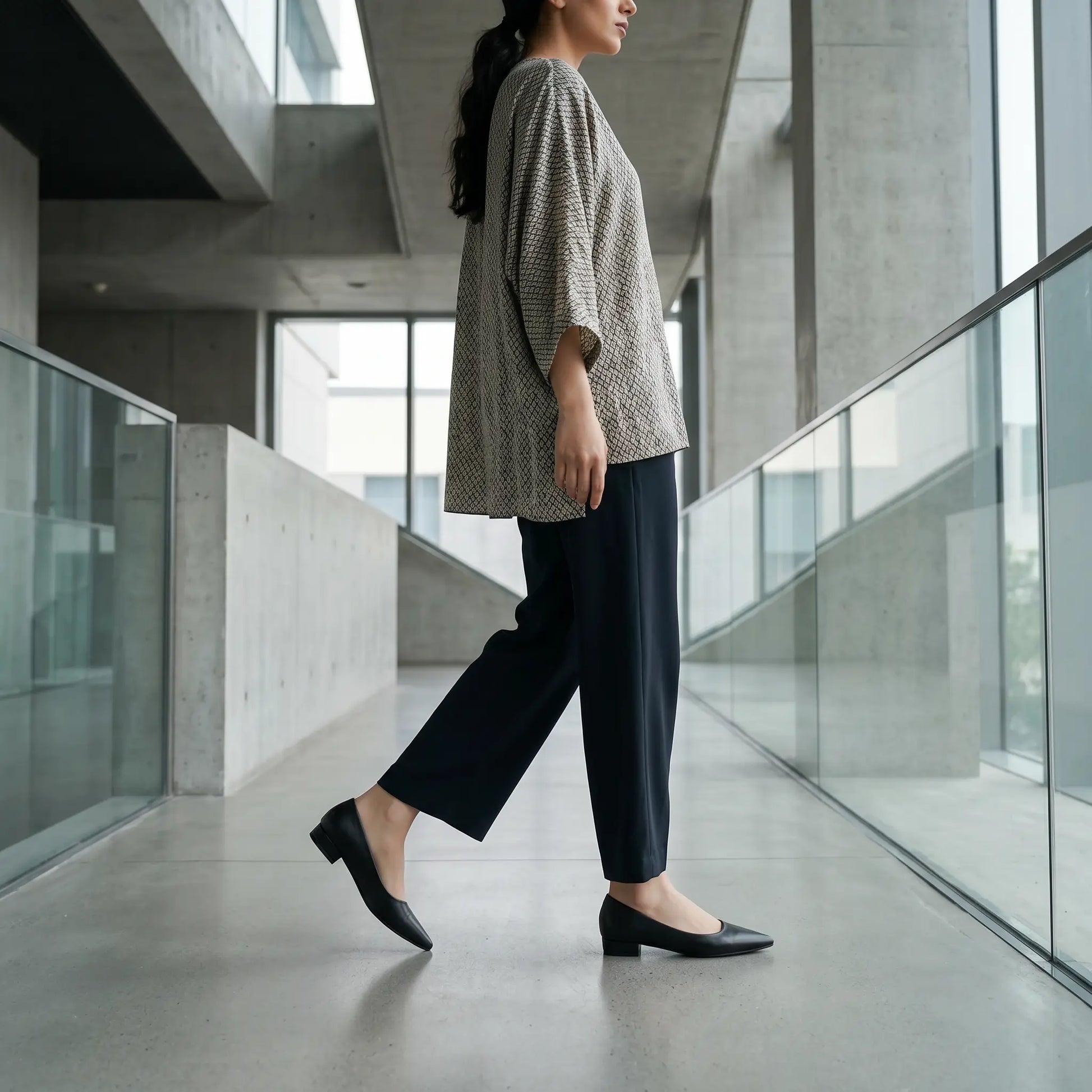 A woman walks forward in black Rhylo pointed-toe flats with a low block heel, styled with wide-leg trousers in a modern architectural setting.