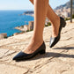 Woman wearing black Rhylo elegant pointed-toe slip-on flats with a low block heel, walking on cobblestones with a scenic coastal city view.