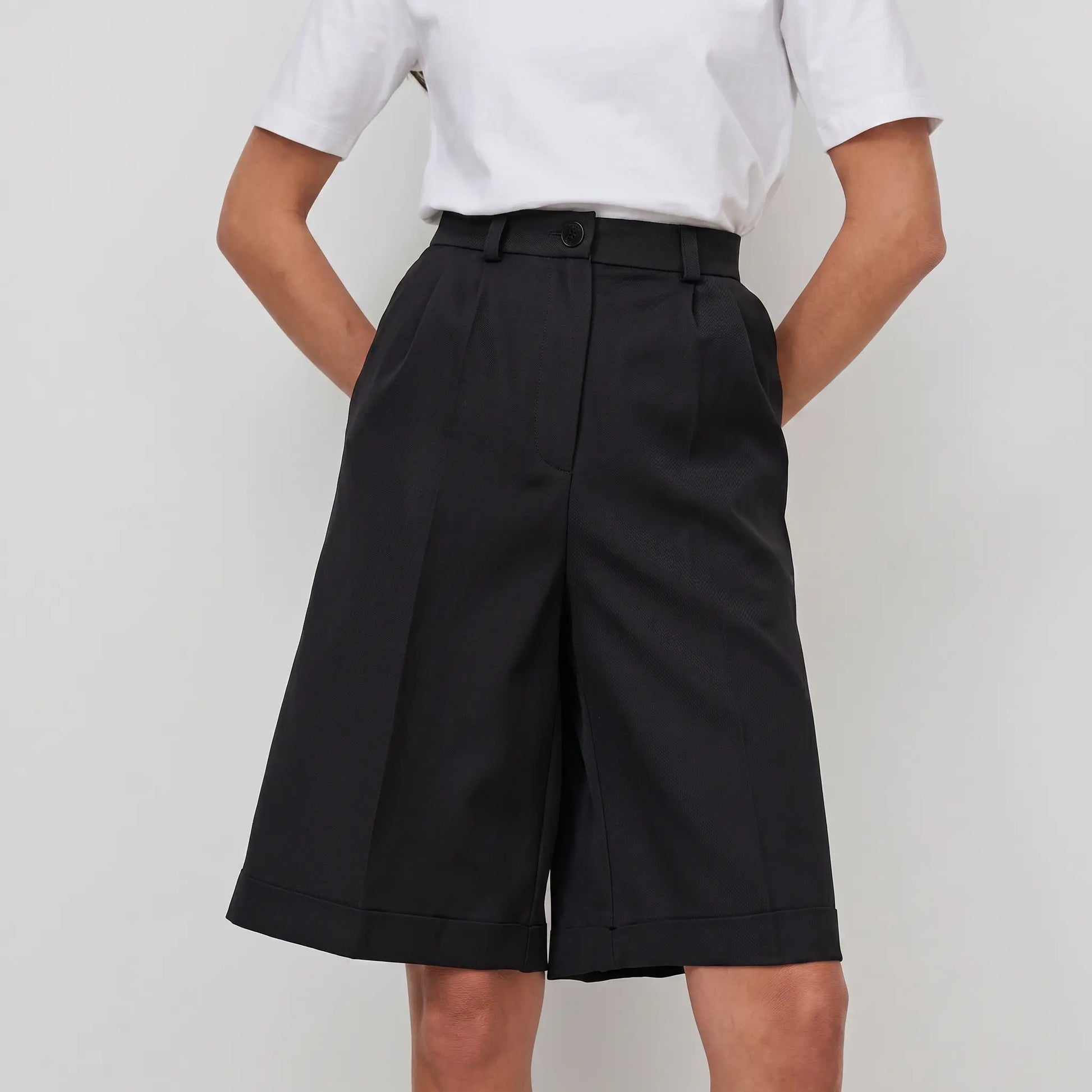 Black high-waisted, wide-leg Bermuda shorts with tailored front pleats and a cuffed hem, shown on a woman against a white studio background.