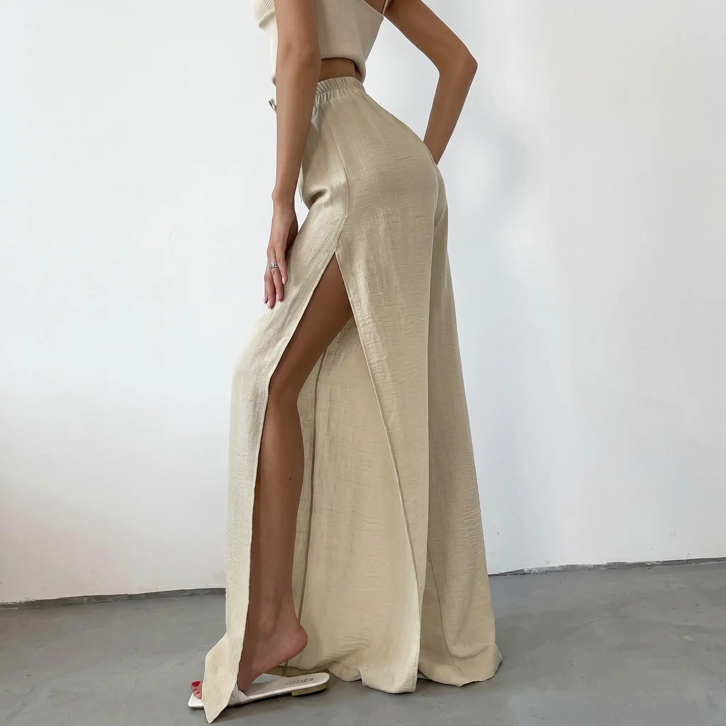 Rhyzen high-waisted wide-leg trousers in a flowing oat beige cotton, featuring dramatic deep side slits. Shown from a three-quarter view on a model in a studio setting.
