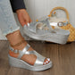 Silver metallic peep-toe wedge sandals with an adjustable ankle strap shown on a model's feet, styled with distressed denim jeans on a wooden floor.