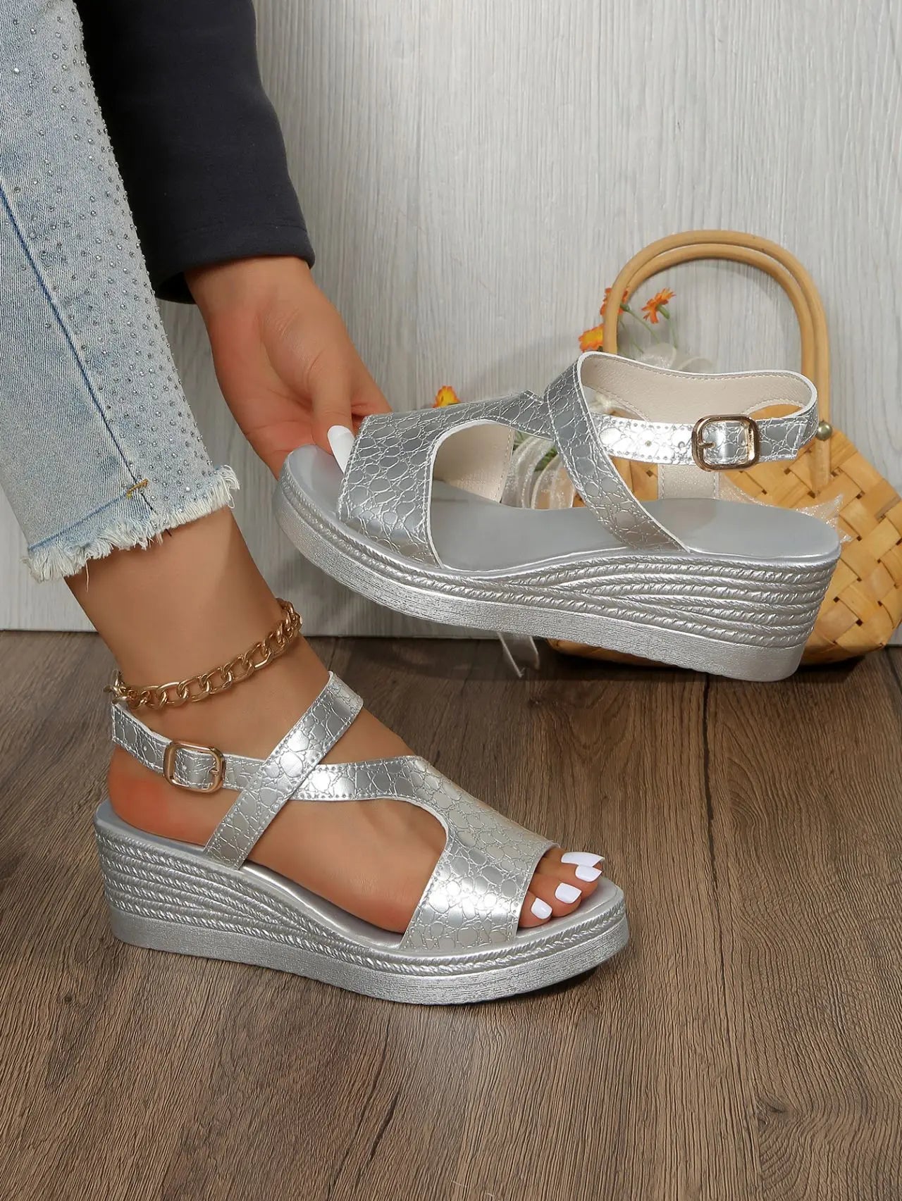 Silver metallic peep-toe wedge sandals with an adjustable ankle strap shown on a model's feet, styled with distressed denim jeans on a wooden floor.