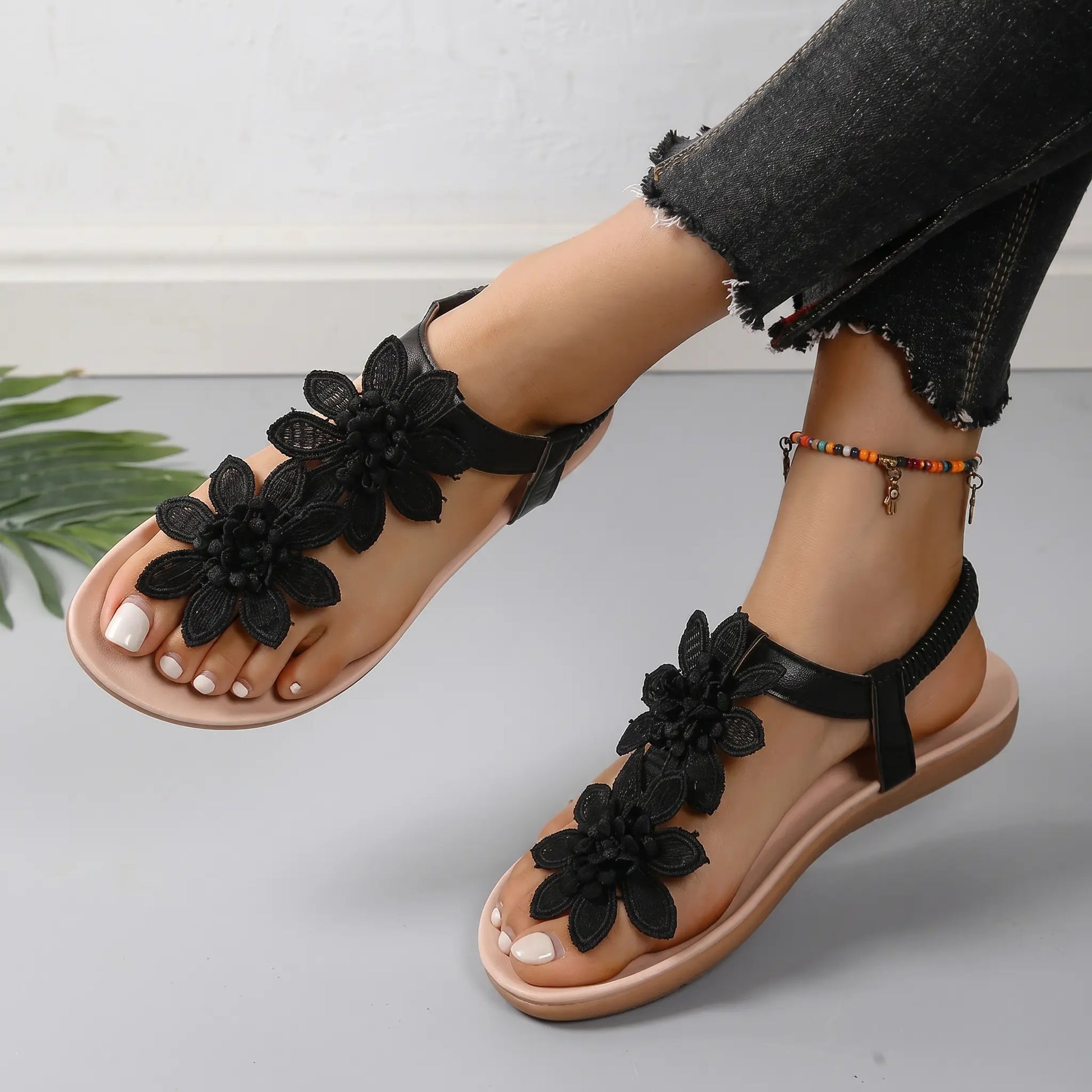 Rishka black bohemian flat sandals adorned with delicate floral appliques on the toe strap and a comfortable elasticated heel strap, showcased on a woman's feet with jeans and a beaded ankle bracelet.