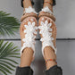 Rishka Bohemian floral embellished white flat sandals worn on feet, featuring delicate lace appliques on the toe strap and a black elastic heel strap. Styled with denim shorts and colorful anklets.