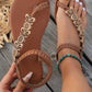 Rivenia bohemian flat sandals in warm brown, featuring hand-woven straps, decorative wood-effect floral and butterfly embellishments, and an elasticized ankle strap, shown worn on a foot with a turquoise anklet.