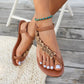 Close-up of Rivenia bohemian woven flat sandals on a woman's feet, featuring intricate braided straps, decorative wood-effect floral embellishments, and an elasticized ankle strap, styled with a turquoise beaded anklet against a wooden deck backdrop.
