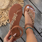 Rivenia bohemian brown woven flat T-strap sandals adorned with carved wood-effect floral and circular embellishments, shown on a model's feet with a turquoise anklet against a wooden deck background.