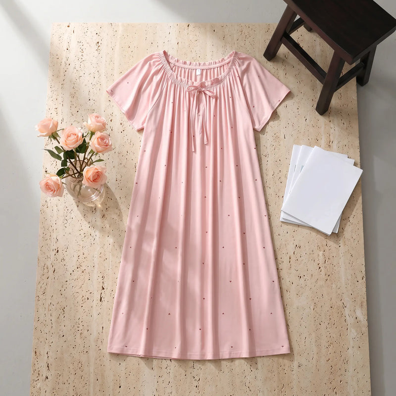 Rosewater Pink Zevryn Romantic Lounge Dress, presented flat-lay, featuring a gathered scoop neckline with a delicate self-tie bow, short flutter sleeves, and a subtle micro-heart pattern, styled on a textured beige surface with pink roses.