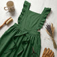 Forest green ruffled cotton pinafore apron with cross-back design, styled with gardening tools and dried flowers on a wooden table.