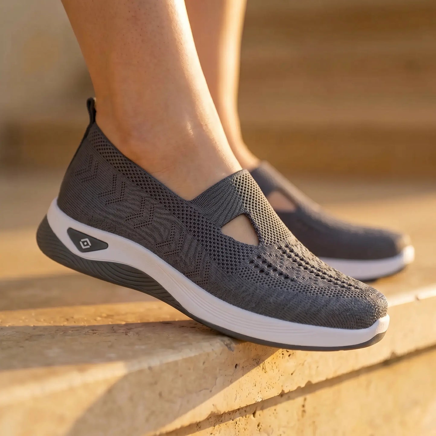 Elegant charcoal Ruxen slip-on sneakers on a model's feet, featuring the breathable woven knit upper, triangular instep cutout, and thick white sole, styled on sunlit stone steps.