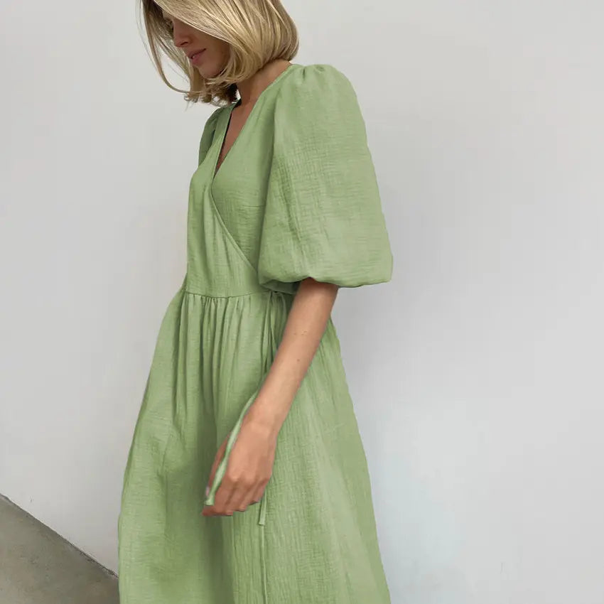 Woman wearing a sage green crinkled cotton gauze midi wrap dress with voluminous short puff sleeves and a V-neckline, shown in profile against a white studio background.