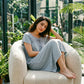 Woman in a Serene Blue Zevryn midi lounge dress, featuring a gathered scoop neck with a delicate bow, flutter sleeves, and a subtle micro-pointelle pattern, seated in a sunlit plant-filled room.