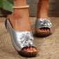 Silver slip-on sandals featuring a delicate floral embellishment and perforated detailing on the strap, showcased on a woman's feet against a textured woven background.