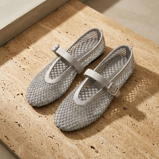 Elegant silver grey Mary Jane flats with crystal mesh upper and diamond netting, featuring a rounded toe and a suede-finish strap with a metal buckle, presented on a textured beige surface.