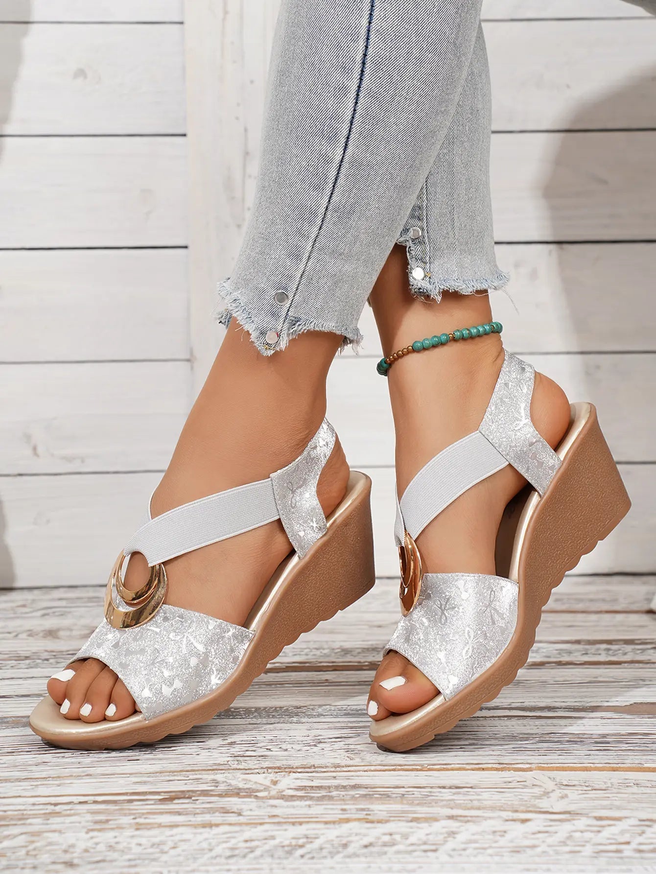 Silver metallic floral print peep-toe wedge sandals with criss-cross elastic straps and a decorative gold-tone ring, worn with casual jeans.