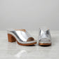 Pair of silver metallic V-cut vamp mules with chunky wood-effect block heels and white contrast stitching, displayed on a marble-like surface.