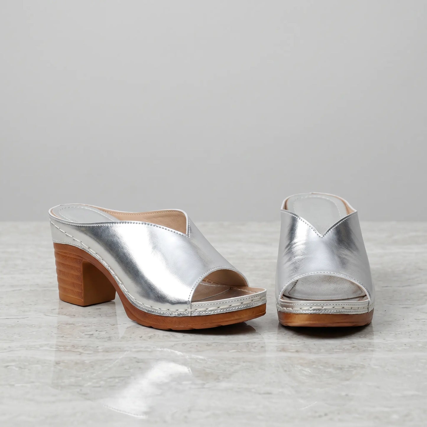 Pair of silver metallic V-cut vamp mules with chunky wood-effect block heels and white contrast stitching, displayed on a marble-like surface.