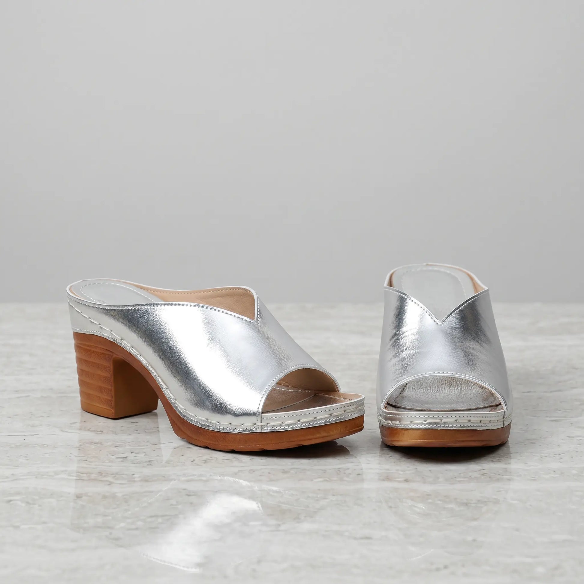 Pair of silver metallic V-cut vamp mules with chunky wood-effect block heels and white contrast stitching, displayed on a marble-like surface.