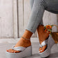 A woman's feet wearing shimmering silver metallic wedge platform flip-flops with a knotted toe strap, styled with embellished jeans against a rustic background.