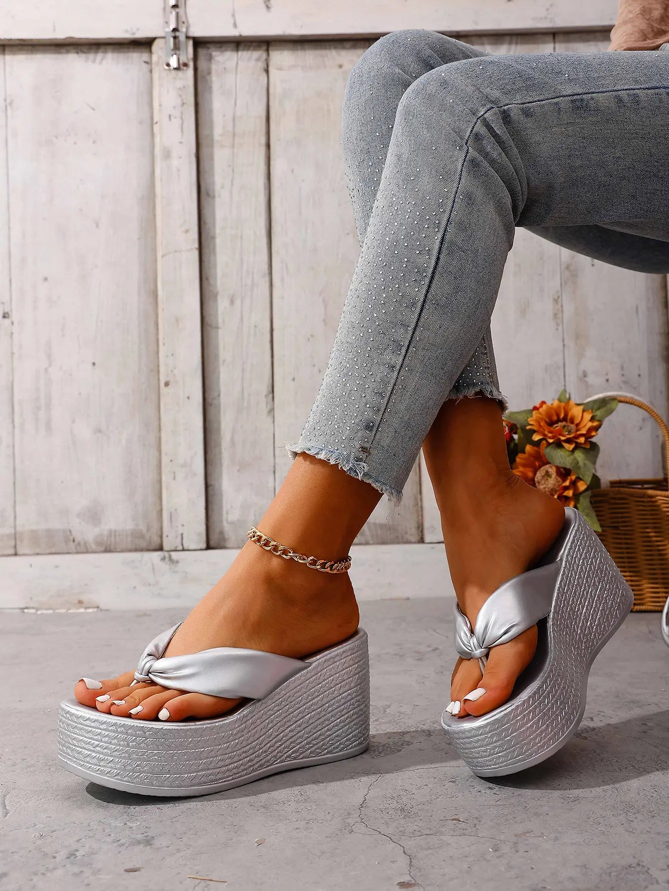 A woman's feet wearing shimmering silver metallic wedge platform flip-flops with a knotted toe strap, styled with embellished jeans against a rustic background.