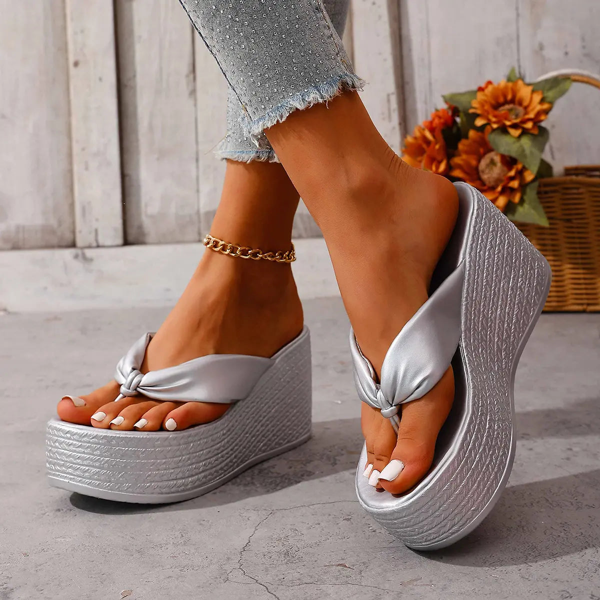 Women's shimmering silver metallic wedge platform flip-flops with a knotted toe strap and woven-textured wedge base, shown on feet from a three-quarter angle with a subtle studio background.