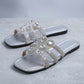 Pair of silver flat slide sandals embellished with sparkling floral rhinestones on a textured studio background.