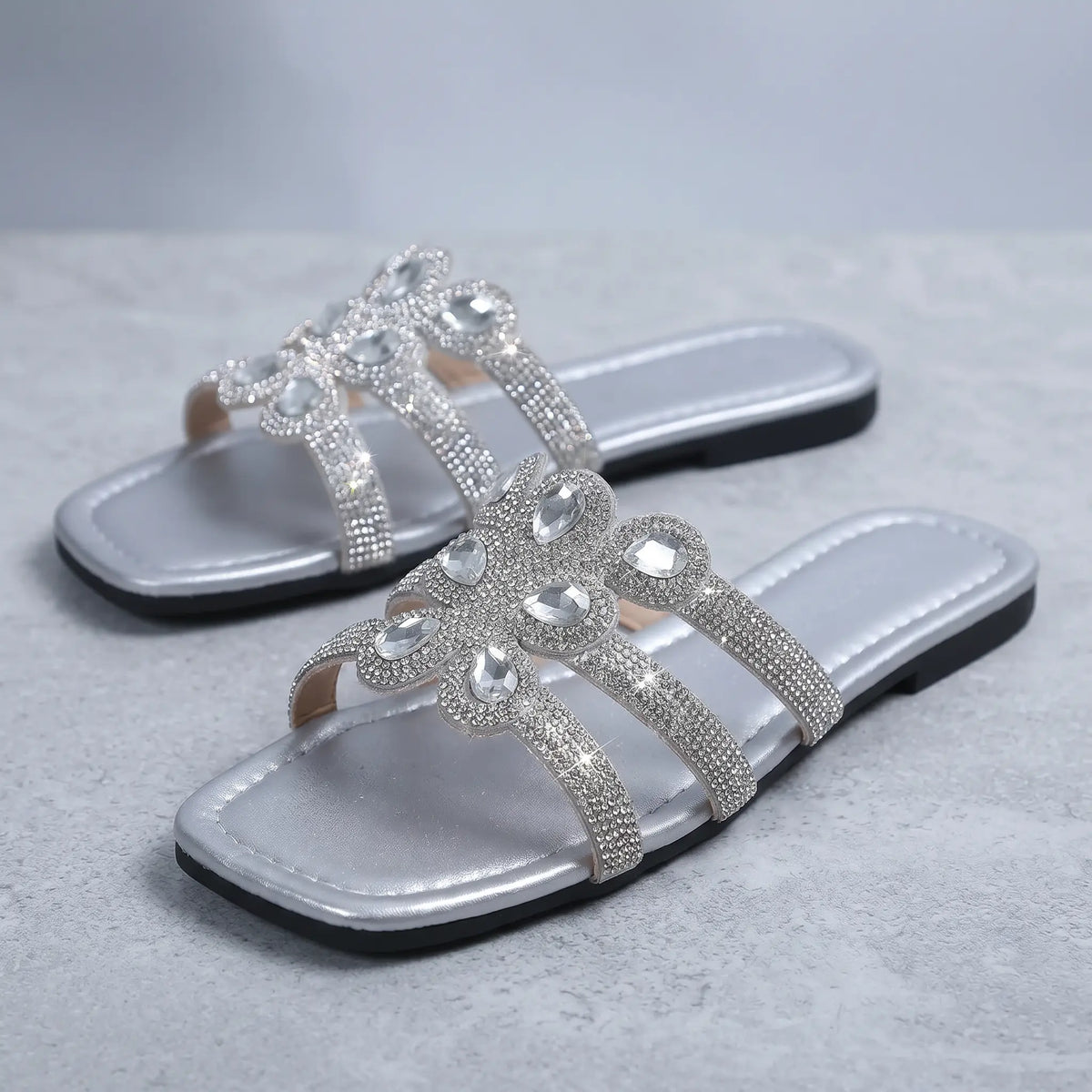 Pair of silver flat slide sandals embellished with sparkling floral rhinestones on a textured studio background.