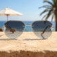 Silver-tone rimless aviator sunglasses with intricate chain-link temple details and gradient grey lenses, set against a blurred beach resort backdrop.