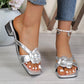 Elegant silver heeled mules adorned with a large fabric rose embellishment, shown on a woman's feet with white-painted toenails, set against a soft lifestyle background with wooden elements. These open-toe slides feature a chunky block heel and square toe for stylish summer wear.