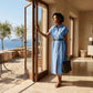A woman in a sky blue linen shirt dress with a black belt stands by a glass door, overlooking a coastal view with olive trees.