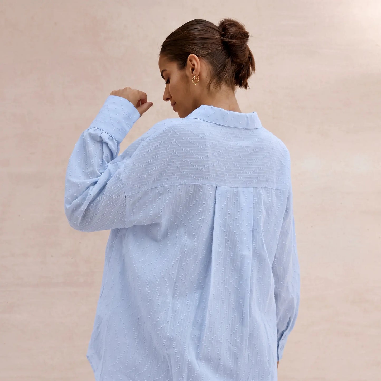Back view of a woman wearing a light sky blue textured cotton oversized shirt with a camp collar and pearl buttons, against a blush pink background.