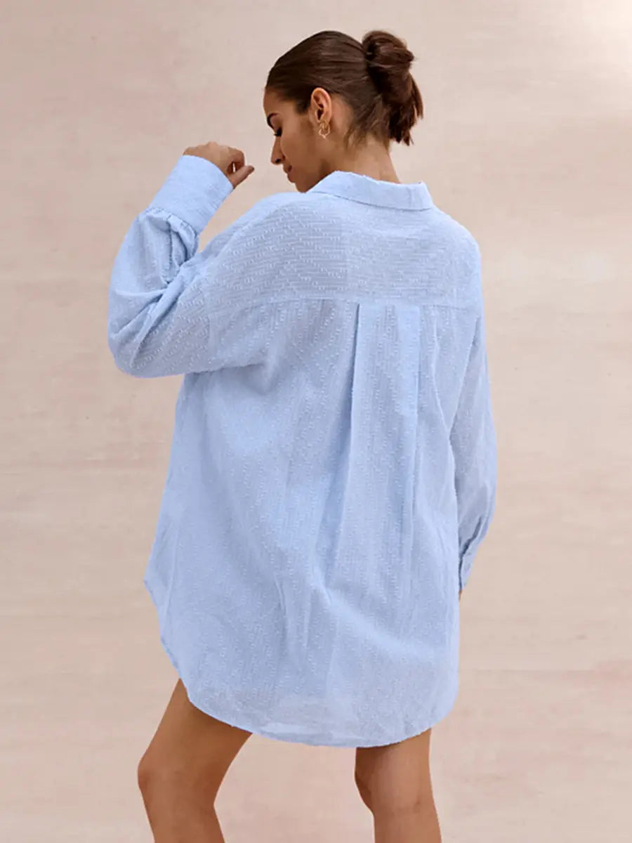 Back view of a woman wearing a light sky blue textured cotton oversized shirt with a camp collar and pearl buttons, against a blush pink background.