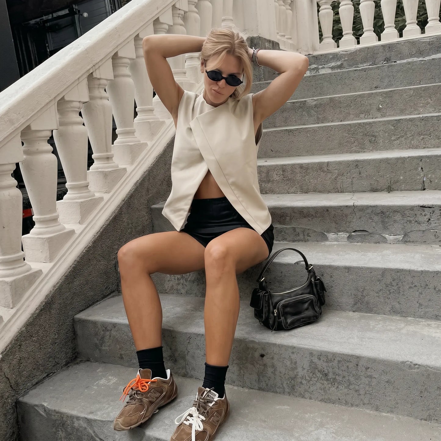 Woman wearing a sleeveless Xylohn asymmetric drape top in champagne beige, styled with black shorts, sunglasses, and jewelry, sitting on outdoor stairs.