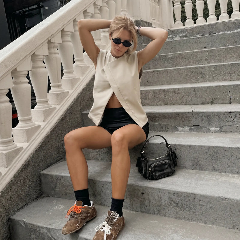 Woman wearing a sleeveless Xylohn asymmetric drape top in champagne beige, styled with black shorts, sunglasses, and jewelry, sitting on outdoor stairs.