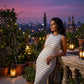 Elegant woman wearing a Zyphron sleeveless ribbed knit maxi dress with off-white and light blue horizontal stripes, leaning on a railing on a twilight rooftop with a city skyline and glowing lanterns.