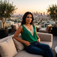 Model wearing a vibrant green sleeveless V-neck blouse with intricate beige crochet trim, seated on a plush sofa on a rooftop with a city skyline at sunset.