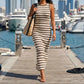Model wears a Zyphron sleeveless ribbed knit maxi dress with sand khaki and off-white horizontal stripes, walking on a wooden pier. The elegant body-contouring dress is accessorized with a straw tote bag, set against a sunny marina background.