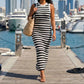 Woman wearing a black and white striped ribbed knit sleeveless maxi dress, walking on a sunny wooden pier with yachts and city buildings in the background. She carries a woven straw bag.