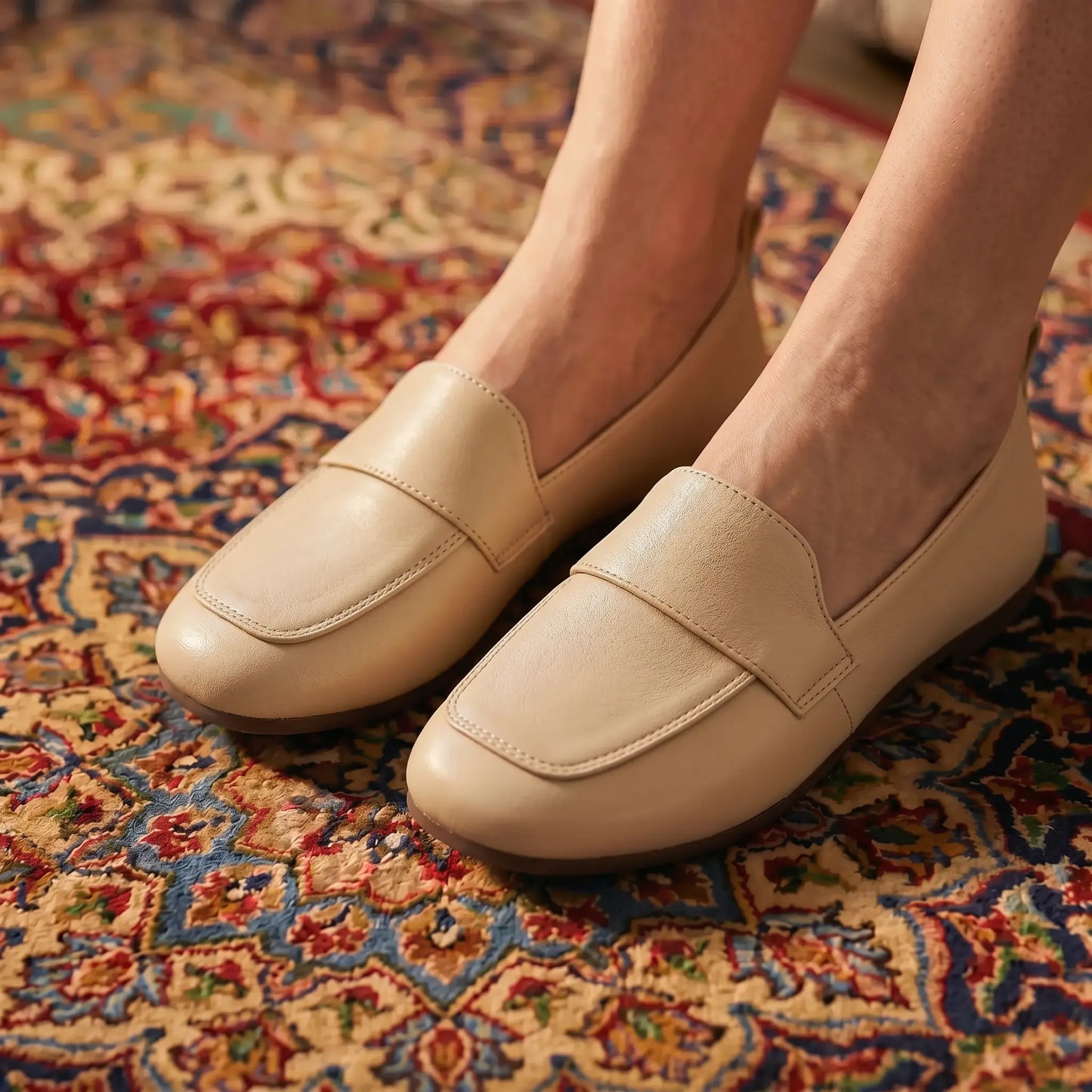 Soft Apricot vegan leather slip-on loafers featuring a classic transverse vamp strap and round toe, worn on a richly patterned rug.