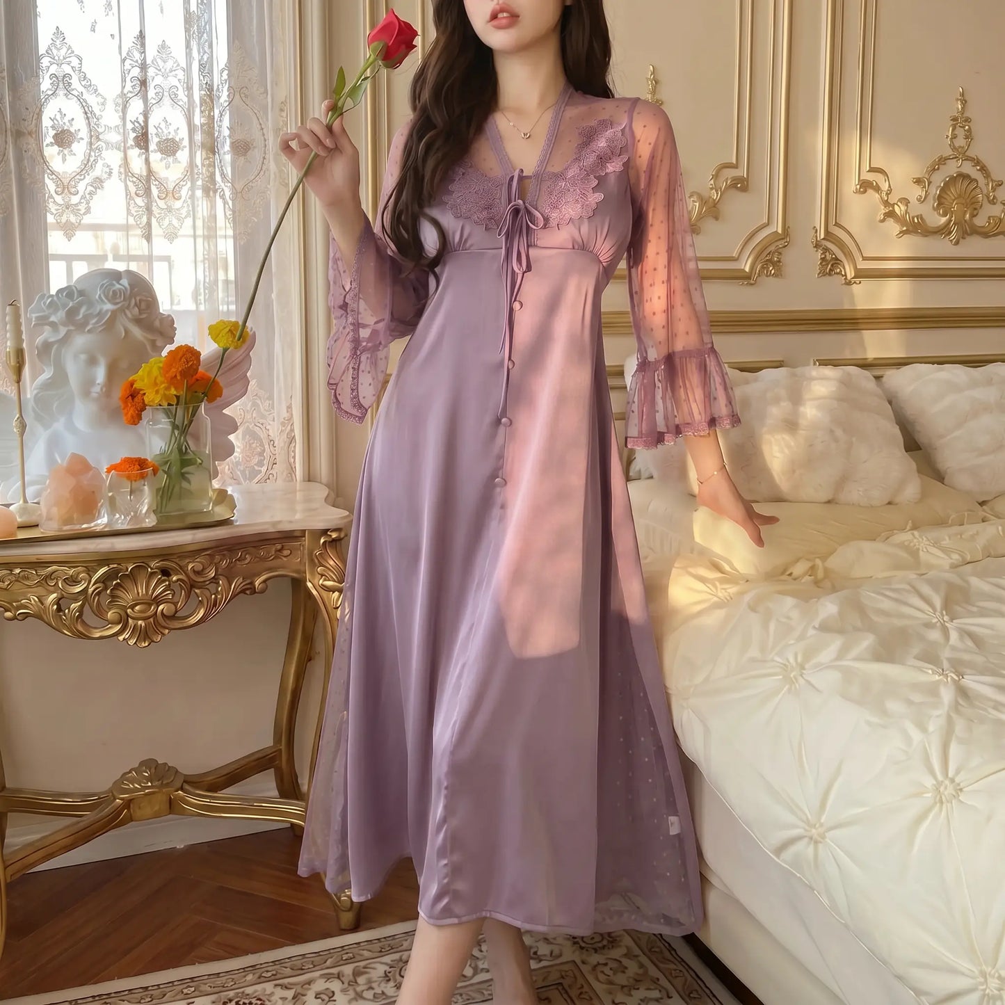 Woman in Soft Lilac Sylven long satin nightgown with floral lace V-neck, front tie, and sheer polka-dot bell sleeves, posing elegantly in a luxurious bedroom holding a red rose.